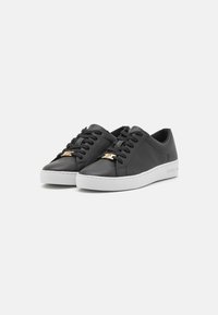 Schwarze Ledersneaker mit weißen Gummisohlen, ausgestattet mit schwarzen Schnürsenkeln und einem goldenen Logo-Akzent auf der Vorderseite. Glatte Textur und abgerundetes Design.