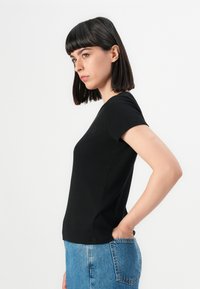 Giovane donna con capelli scuri lisci, che indossa una maglietta nera a maniche corte e jeans blu, in piedi di profilo con una mano nella tasca posteriore.