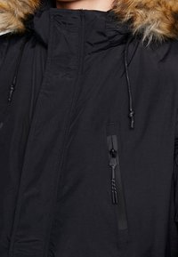 Svart isolerad parka med huva klädd i fuskpäls, snoddar och dragkedjefickor. Slät tyg med matt finish.