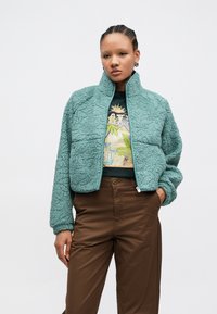 Miętowa, teksturowana kurtka polarowa z zamkiem z przodu, wysokim kołnierzem i przednimi kieszeniami; noszona na koszulce z nadrukiem i brązowych spodniach.