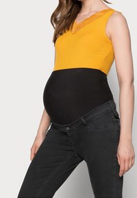 Těhotná žena v žlutém bezrukávovém topu a černých těhotenských džínách s elastickým pásem, stojící na jednobarevném pozadí.