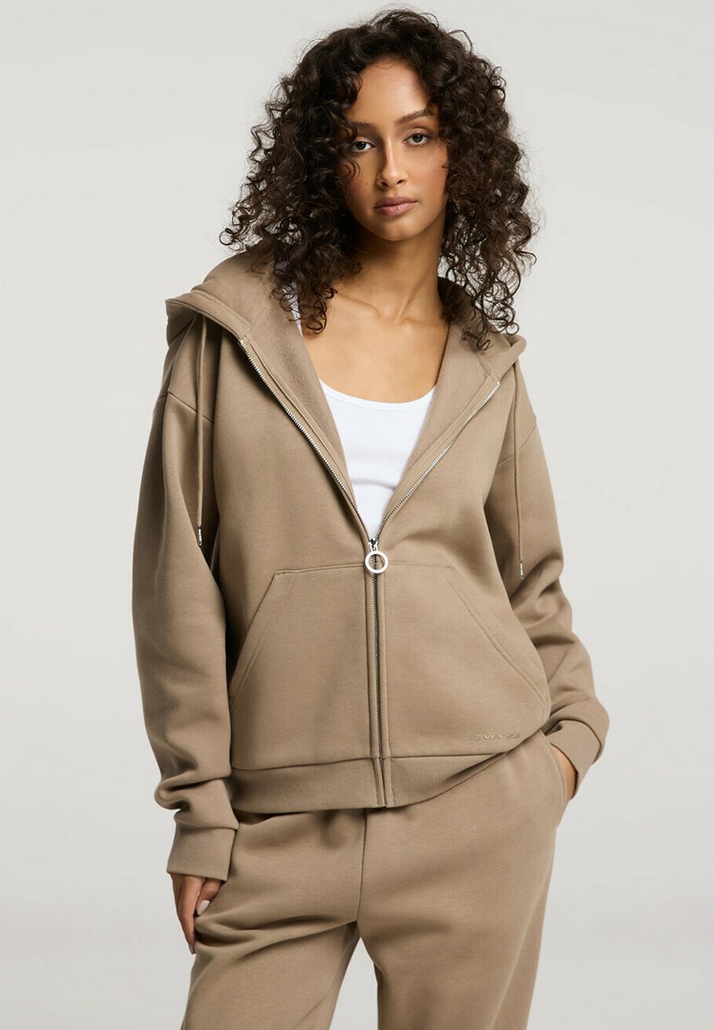 Beżowa bluza z kapturem na zamek, wykonana z miękkiego materiału, z dużym kapturem, kieszeniami z przodu i ściągaczami na rękawach. Połączona z pasującymi spodniami.