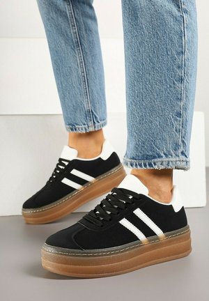 Personne portant des baskets plateforme noires et blanches aux semelles en gomme et un jean court bleu clair, debout sur un sol gris.