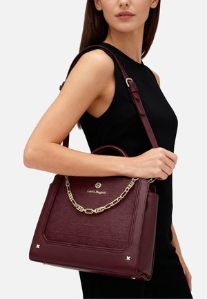 Donna che indossa un vestito nero senza maniche, tiene una borsa in pelle color bordeaux con catena e logo dorati su sfondo bianco.