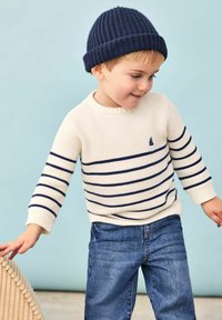 Gestickter cremefarbener Pullover mit marineblauen Streifen und kleinem Segelboot-Logo, kombiniert mit blauen Jeans und einer marineblauen Rippmütze, vor einem hellen Hintergrund.