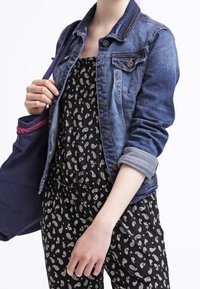 Chaqueta de mezclilla en azul medio con detalles de botones y puños enrollados, usada sobre un mono negro con un patrón de paisley blanco.