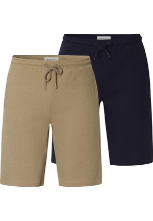 Mann trägt beige, strukturierte Shorts mit Kordelzug, ein dunkelblaues Hemd und schwarze Sneaker mit weißen Sohlen auf einfarbigem Hintergrund.