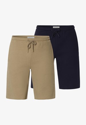 Mann trägt beige, strukturierte Shorts mit Kordelzug, ein dunkelblaues Hemd und schwarze Sneaker mit weißen Sohlen auf einfarbigem Hintergrund.