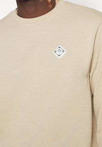 Gros plan d'une personne portant un t-shirt beige à col rond avec un petit patch logo carré blanc sur la poitrine gauche.