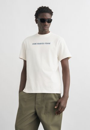 Férfi fekete napszemüvegben, rövid ujjú fehér pólóban a "The North Face" logóval és olívazöld nadrágban, egyszínű háttér előtt állva.