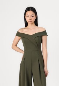 Tuta verde oliva scollata con corpino aderente e design a gamba larga plissettata. Tessuto liscio e dettagli di cucitura sottili.