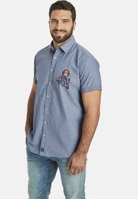 Hellblaues Kurzarmhemd mit Knopfkragen, verziert mit gestickten Details und einem Etikett am Saum, kombiniert mit hellblauen Jeans.