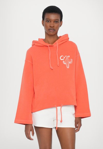 Korallenfarbener, übergroßer Hoodie mit Kordelzügen, versehen mit einem Kaktus-Graphic und dem Text 'Closed'. Kombiniert mit weißen Shorts, aus einem glatten Baumwollstoff.