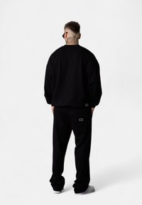 Man with short hair and back neck tattoo wearing black sweatshirt, black pants, white sneakers, and black sunglasses, standing facing away.