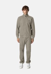 Giacca in pile grigio chiaro con zip e colletto alto, abbinata a pantaloni tuta coordinati. Presenta polsini a coste e un logo sul petto.