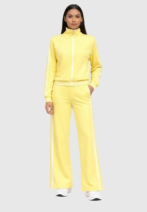 Femme debout portant une veste de sport jaune à fermeture éclair et un pantalon large assorti avec des bandes blanches, ainsi que des baskets blanches.