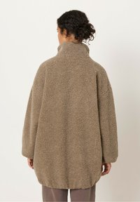 Ruskea pehmeä fleece-takki, jossa on korkea kaulus ja ylisuuri istuvuus. Ominaisuuksiin kuuluu pyöristetty helma ja väljät hihat lisäämään mukavuutta.