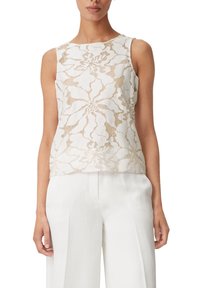 Ärmlös beige spetsblus med stora blommönster, bärs med högmidjade vita vida byxor på en stående modell.