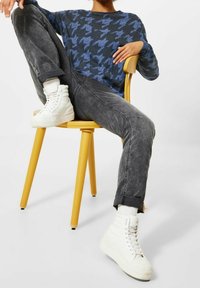 Blå och svart houndstooth-mönstrad tröja, grå jeans med uppvikta anklar och vita högstövlar, sittande på en gul stol.