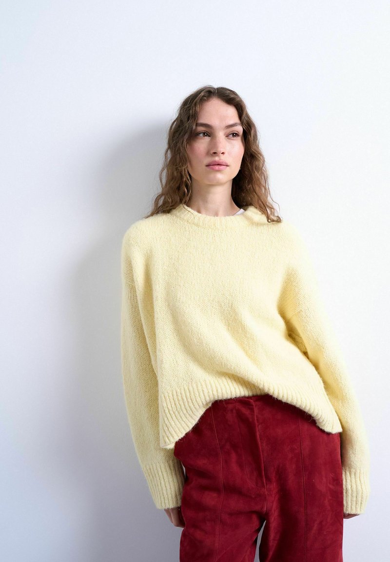 Jeune femme aux cheveux ondulés portant un pull ample jaune pâle et un pantalon rouge texturé, debout contre un mur blanc.