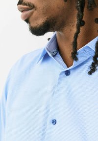 Svetlomodrá textúrovaná košeľa s vnútorným golierom s potlačou, vybavená zapínaním na gombíky vpredu a priliehavým strihom. Výrazný detail s modrým gombíkom.