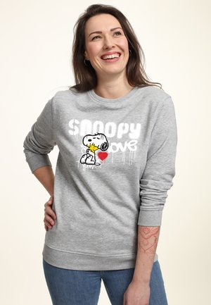 Smilende kvinde med langt brunt hår iført grå Snoopy-sweatshirt, der holder et rødt hjerte, står med hånden på hoften og har en tatovering på underarmen.