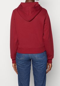 Sudadera burdeos con bolsillo frontal y capucha con cordón. Hecha de una suave mezcla de algodón, con puños y dobladillo acanalados, combinada con jeans azules.