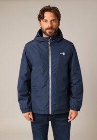 Veste imperméable marine avec capuche, fermeture éclair, poignets élastiques et deux poches latérales. Présente un patch logo sur la poitrine gauche.