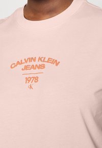 Růžové bavlněné tričko s kulatým výstřihem. Uprostřed se nachází logo v oranžové barvě, které zní "CALVIN KLEIN JEANS 1978" s malým akcentem "ck" níže.