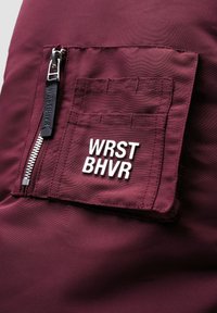 Burgundy tyg med en ficka med dragkedja, en rektangulär applikation med "WRST BHVR" i vitt och en slät, texturerad yta.