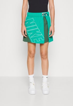 Γυναίκα φορώντας μια πράσινη φούστα Nike με μεγάλο λογότυπο, λευκές κάλτσες Nike και λευκά sneakers, στέκεται μπροστά σε έναν απλό λευκό φόντο.