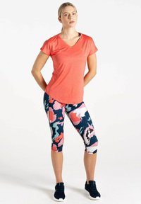 Camiseta de manga corta color coral con escote en V, combinada con leggins azul marino que presentan coloridos patrones florales abstractos. Se llevan deportivas negras.