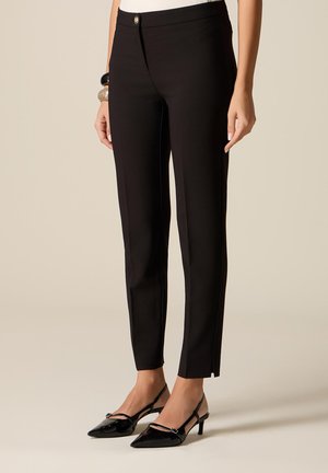 Kvinde iført tætsiddende sorte ankellange bukser med frontfold og sorte spidse slingback-hæle.