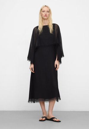 Blonde Frau, die ein durchsichtiges schwarzes Midi-Kleid mit langen Ärmeln und Fransen am Saum sowie schwarze Sandalen mit offenen Zehen trägt, steht vor einem weißen Hintergrund.