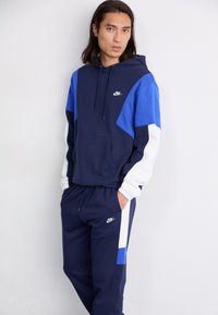 Sötétkék kapucnis pulcsi és melegítőnadrág szett, kontrasztos kék és fehér panelek, húzózsinóros kapucni, valamint Nike logó a mellrészen és a nadrágon.