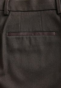 Un pantalon en tissu marron foncé présente une finition texturée, des plis sur les côtés et une poche arrière unique avec un bord contrastant.