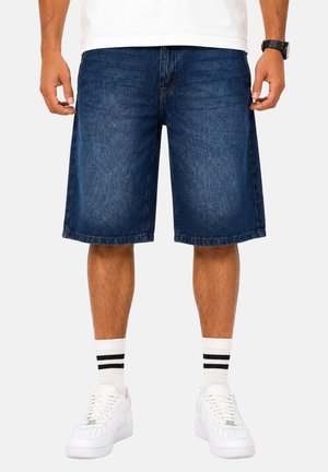 Mann, der dunkelblaue Jeans-Shorts, weiße Socken mit zwei schwarzen Streifen, weiße Sneakers und ein weißes Hemd trägt und mit entspannten Armen steht.