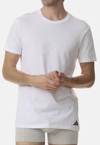 Biała bawełniana koszulka z krótkimi rękawami i okrągłym dekoltem. Posiada czarne logo Adidas na dolnym brzegu. Gładka struktura, luźny krój.