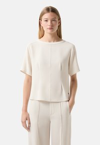 Kurzärmlige, cremefarbene Bluse mit geradem Schnitt, rundem Ausschnitt, sichtbaren Nähten und minimalistischer Gestaltung. Weicher, glatter Stoff.