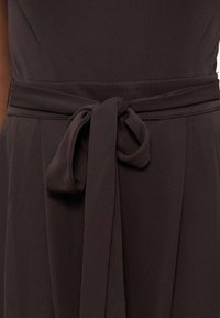 Ζώνη από σκούρο καφέ ύφασμα δεμένη σε φιόγκο γύρω από τη μέση ενός ομοιόμορφου ενδύματος με πιετίτες λεπτομέρειες κάτω από τη ζώνη.