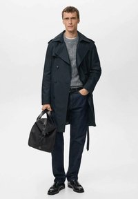 Abrigo oscuro tipo gabardina con solapas grandes y botones, suéter gris de punto, jeans oscuros, zapatos de cuero negros y un bolso negro. Diseño minimalista.