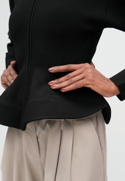 Crna uska jakna s peplum rubom i dugim rukavima, uparena sa svijetlosmeškim plisiranim hlačama. Tekstuirana tkanina, suptilni detalji šavova.
