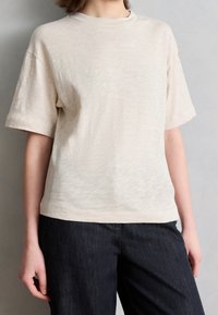 Personne portant un t-shirt uni beige clair à manches courtes associé à un jean foncé, debout devant un fond gris neutre.
