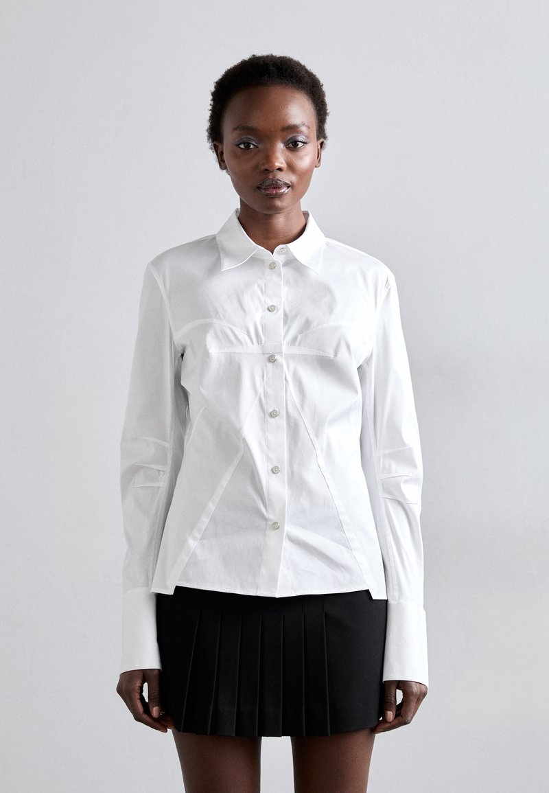 White button-up shirt with structured seams, long sleeves, and pointed collar; paired with a black pleated mini skirt. Smooth fabric texture.