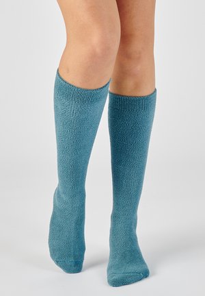 Jambes portant des chaussettes texturées turquoise jusqu'aux genoux, debout sur un fond gris clair uni.