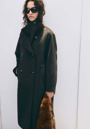 Femme portant un manteau sombre oversize et des lunettes de soleil, tenant un sac marron en fourrure, debout devant un fond blanc uni.