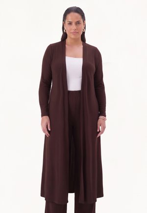 Vrouw met donker haar die gouden oorringen draagt, een witte top, en een lange donkerbruine open cardigan en bijpassende broek tegen een witte achtergrond.