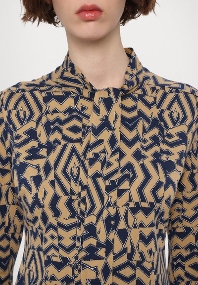 Patterned blouse in beige with navy abstract geometric design. Features a high collar and button placket with subtle texture.