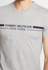 Szara bawełniana koszulka z nadrukiem "TOMMY HILFIGER" i "NOWY JORK" w kolorze granatowym i białym, z poziomymi paskami poniżej tekstu.