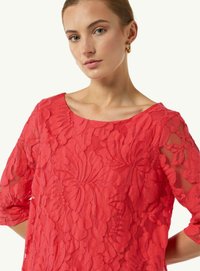 Rotes Spitzentop mit floralen Mustern, weitem Ausschnitt und Dreiviertelärmeln. Die Textur ist detailliert mit einer durchsichtigen Überlagerung.
